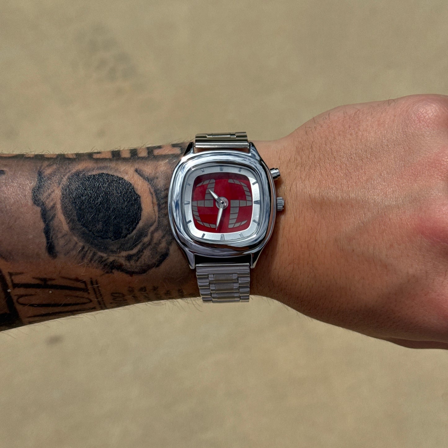 Silver watch with a red face on a wrist with tattoos against a beige background