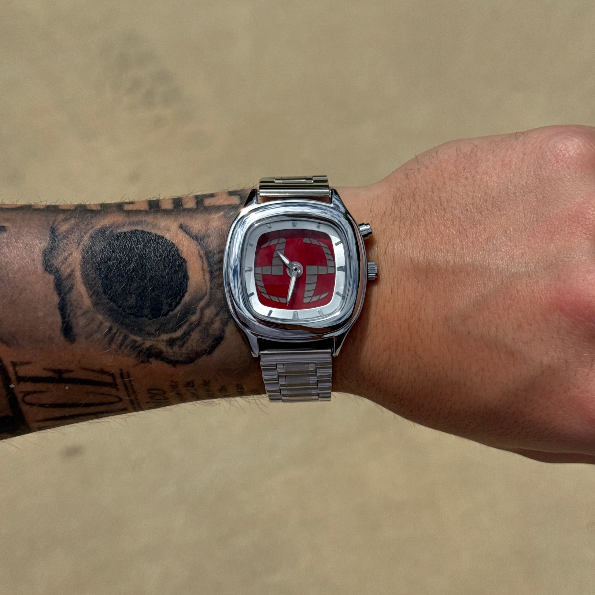 Silver watch with a red face on a wrist with tattoos against a beige background